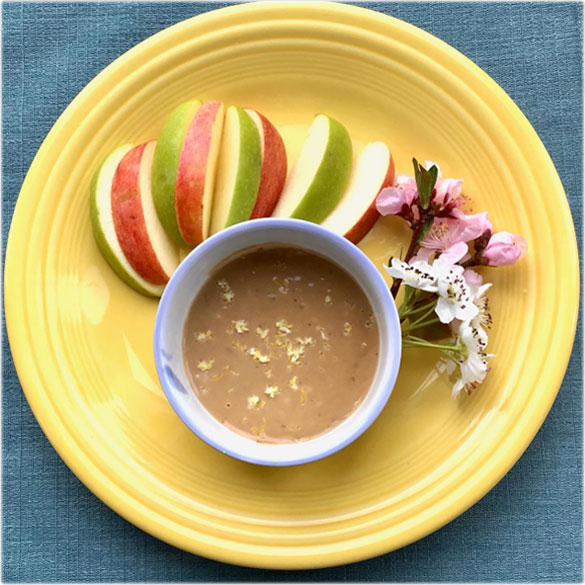 Tahini dip in a bowl with apple slices and flowers on a yellow plate