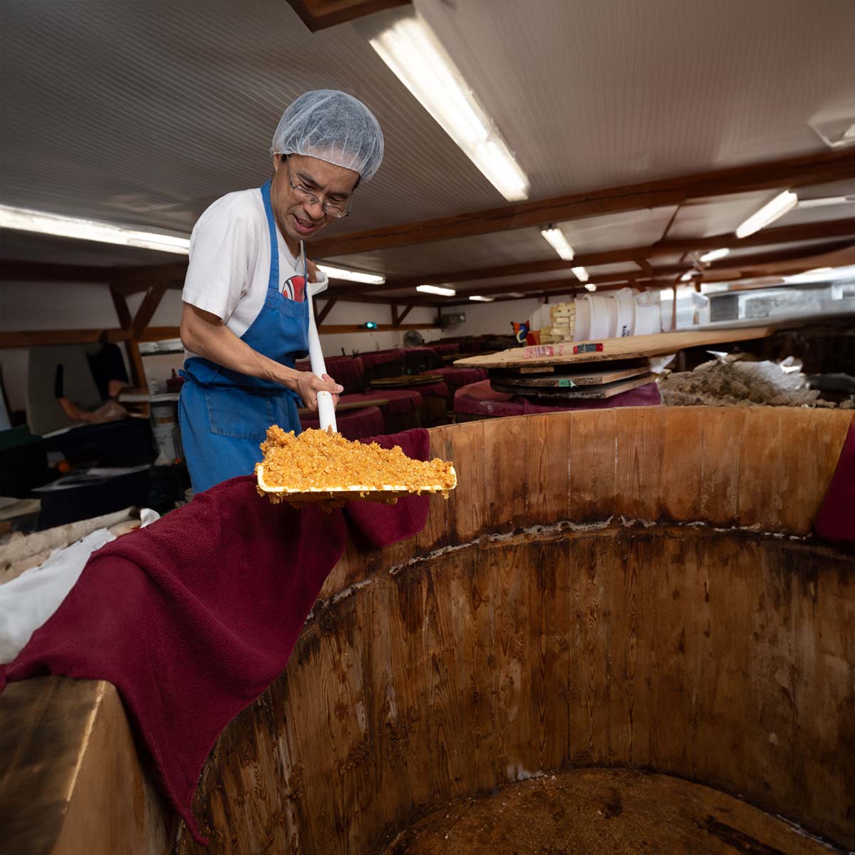 Worker scooping miso out of a large wooden vat and inspecting it